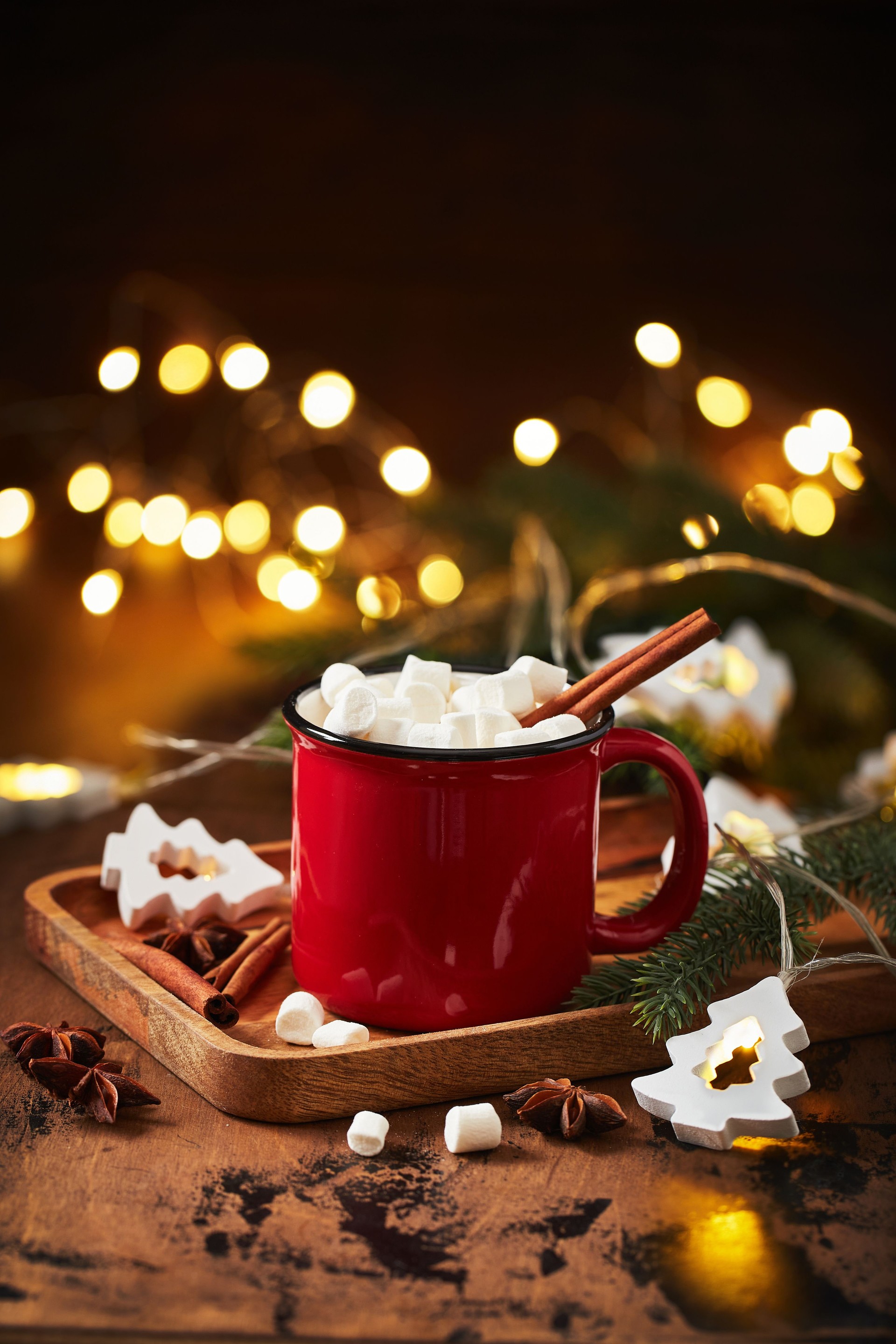 Red cup of hot cocoa with marshmallows on wooden table with christmas tree and glowing garland for christmas