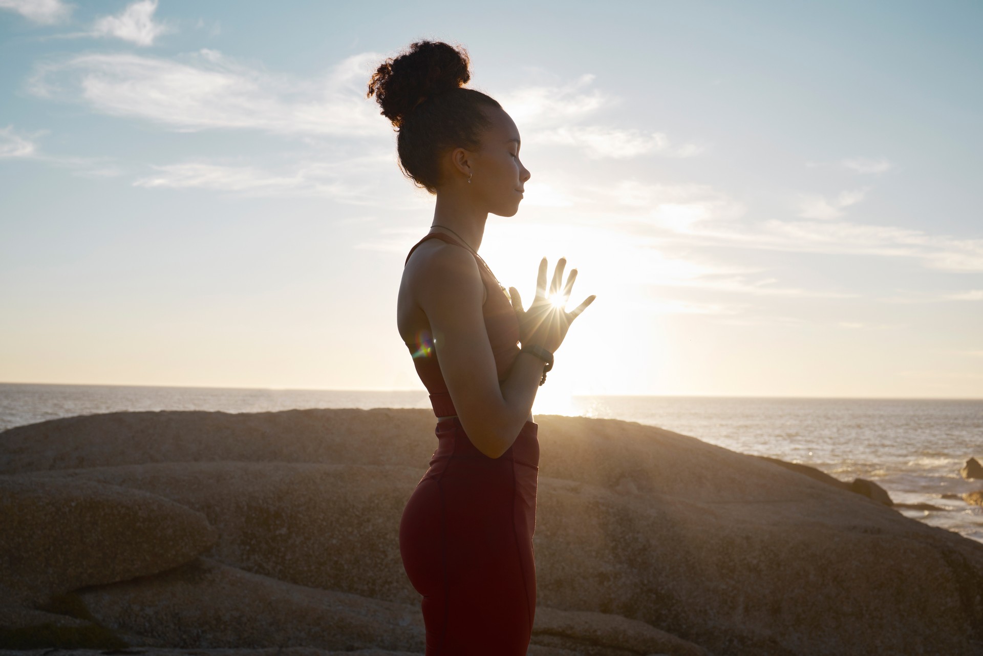 Wellness, yoga and meditation with black woman and fitness at the beach, calm and peace in the sun with lens flare. Zen, mindfulness and healthy mindset, athlete on rocks by the ocean for exercise.