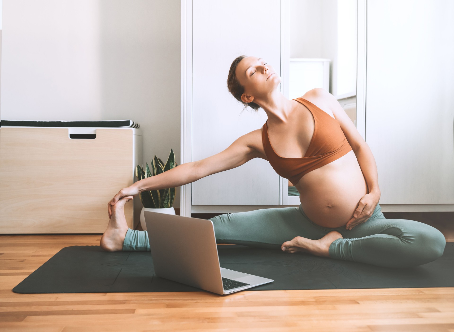 Pregnant woman practicing yoga at home with laptop. Expectant mother doing prenatal video training class indoors.